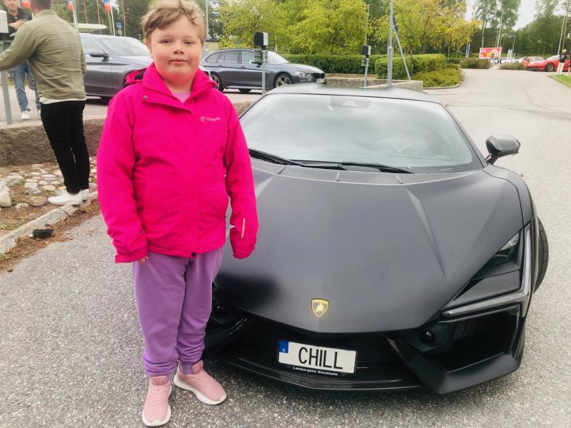 Ett barn i en ljusrosa jacka står bredvid en mattsvart Lamborghini med registreringsskylten