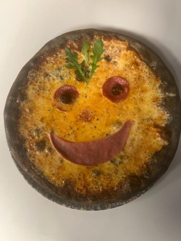 Pizza med svärtad botten, arrangerad för att likna ett leende ansikte med skinka, pepperoni, oliver och en bit ruccola som dekoration.