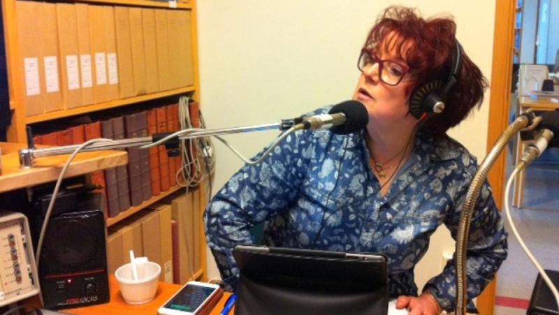 En person med hörlurar talar i en mikrofon i en inspelningsstudio, med bokhyllor, papper och ljudutrustning synliga omkring sig.
