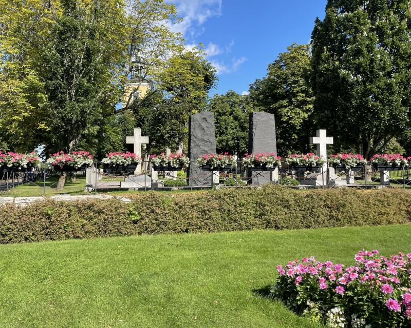 En välskött kyrkogård med två stenkors, två upprättstående stenplattor, blomsterarrangemang och grönt gräs under en klarblå himmel.