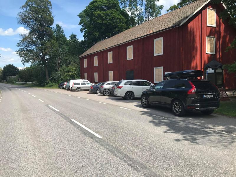 En rad med bilar parkerade längs en vägkant bredvid en stor röd lada under klar himmel.