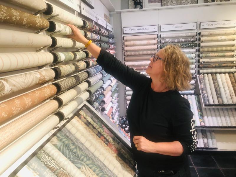 En person står i en butik och sträcker sig efter en rulle mönstrad tapet bland många som visas på hyllorna.