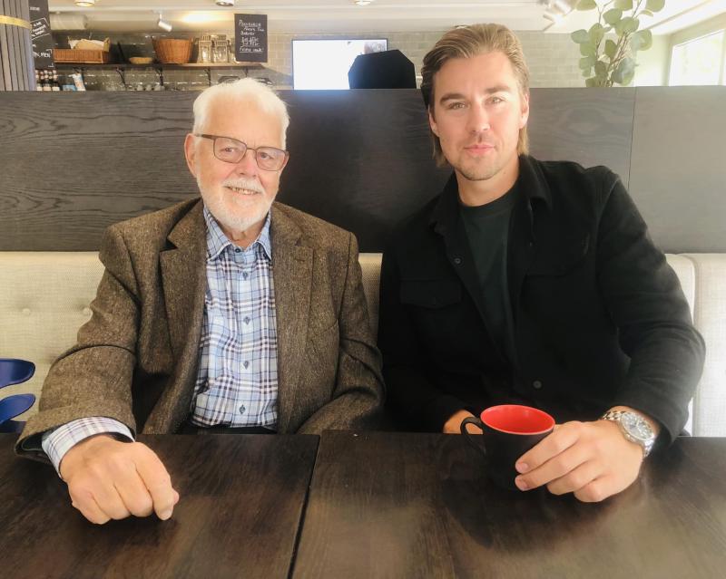 En äldre man och en yngre man sitter tillsammans vid ett bord på ett kafé, båda ler. Den äldre mannen bär glasögon och en rutig skjorta under en kavaj; den yngre mannen håller en svart mugg med röd insida.