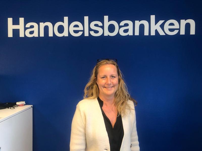 En person som står framför en blå vägg med Handelsbankens logga. De ler och bär en vit jacka över en svart skjorta.