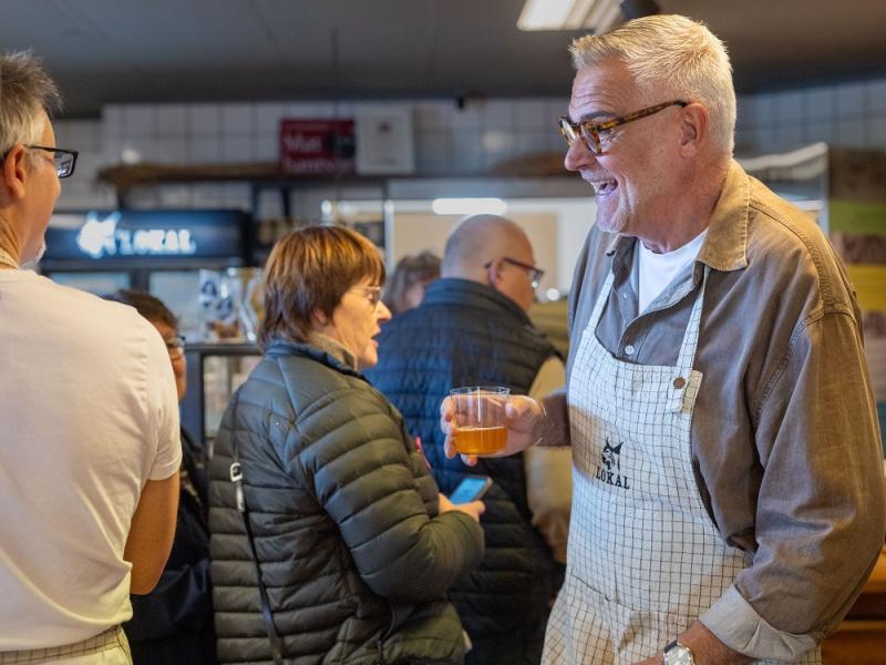 En man med glasögon och förkläde håller i en drink och ler medan han pratar med människor i en livlig inomhusmiljö.