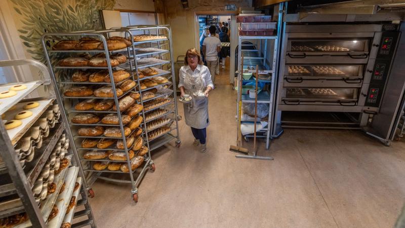 En kvinna i förkläde bär en bricka i ett kommersiellt bageri, omgivet av brödställ och industriugnar.