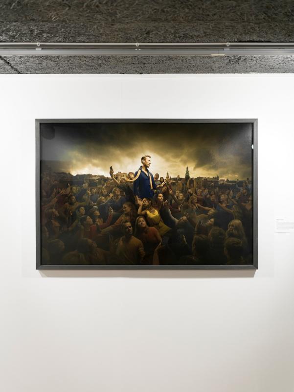 En inramad målning föreställer en skara människor som sträcker sig mot en central figur som står upphöjd ovanför dem, utställd i ett galleri med vit väggbakgrund.