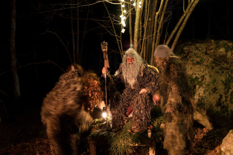Tre personer i dräkter som liknar mossa eller lövverk samlas utomhus på kvällen; en sitter på en träsits och håller i en stav, upplyst av lampor och omgiven av träd.