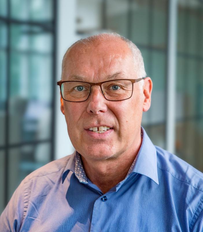 En medelålders man med kort, grått hår och glasögon är klädd i en blå skjorta och står framför en suddig kontorsbakgrund.