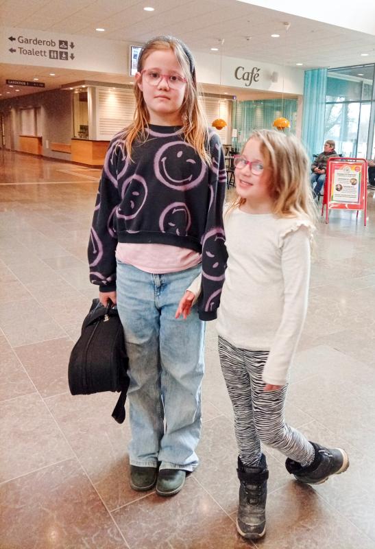Två unga flickor står inomhus; den ena håller i en svart väska och bär glasögon, en smiley-tröja och jeans, medan den andra bär glasögon, en ljus topp och randiga leggings.