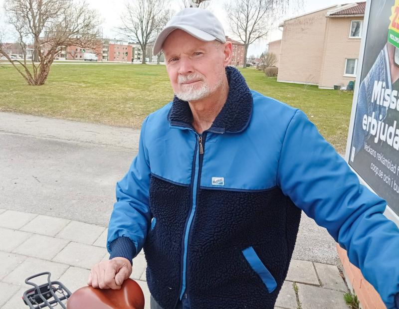 En äldre man i blå jacka och grå keps står utomhus bredvid en cykel på en trottoar, med gräs, träd och byggnader i bakgrunden.
