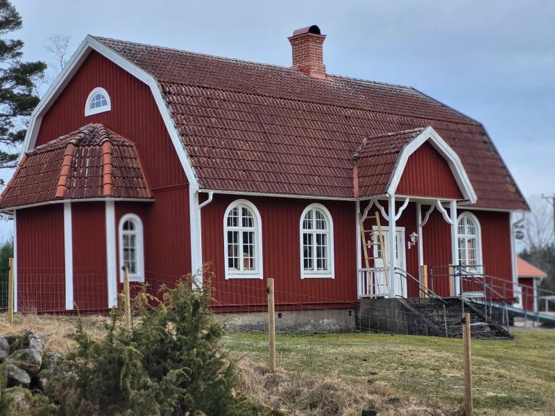 Ett rött trähus med vita detaljer, välvda fönster, tegeltak och en liten veranda ligger på en gräsmatta med staket.