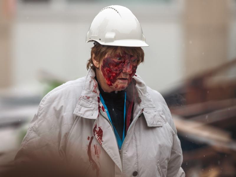 En skadad person iförd vit skyddshjälm och rock med blod i ansiktet och på kläderna står utomhus i närheten av bråte.