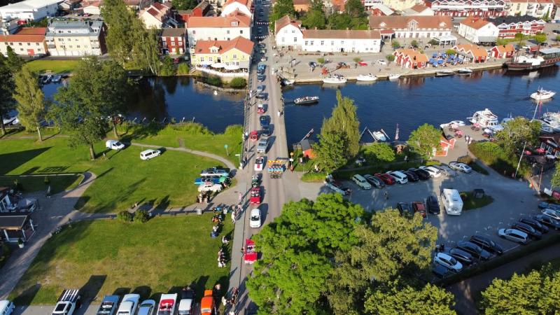 Flygfoto över en stad vid floden med parkerade bilar, människor som går längs en bro, båtar i vattnet och grönområden på båda sidor av floden.