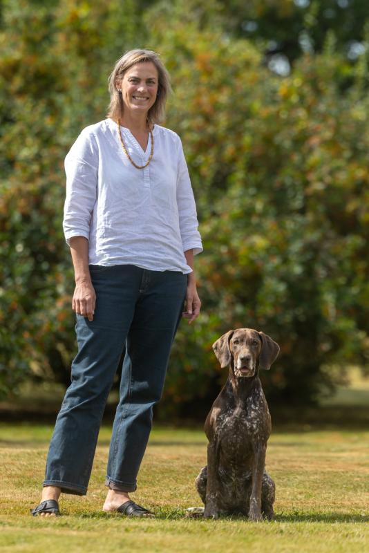 En kvinna i vit skjorta och jeans står på gräset bredvid en sittande brun och vit hund, med gröna buskar i bakgrunden.