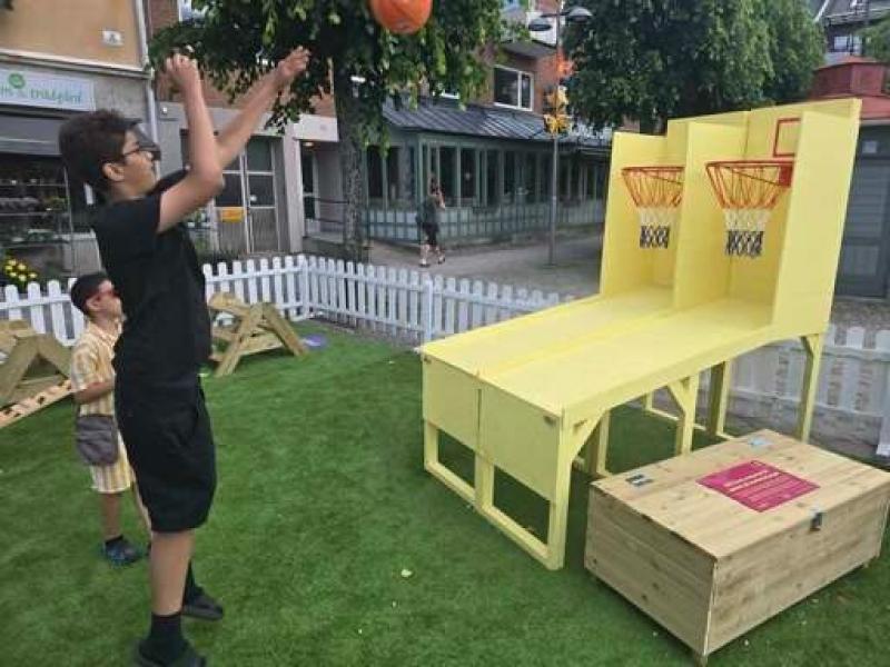 Två pojkar spelar ett minibasket-arkadspel utomhus på en gräsplan med ett trästaket och byggnader i bakgrunden.