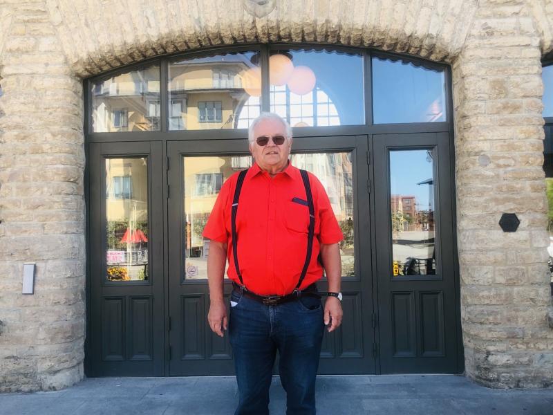 En äldre man i ljusröd skjorta med hängslen står framför en byggnad med välvda stenar och speglande glasdörrar.