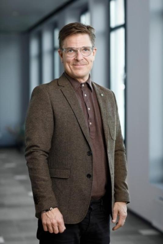 En man med glasögon, en brun kavaj och en brun pikétröja står i en korridor med stora fönster.