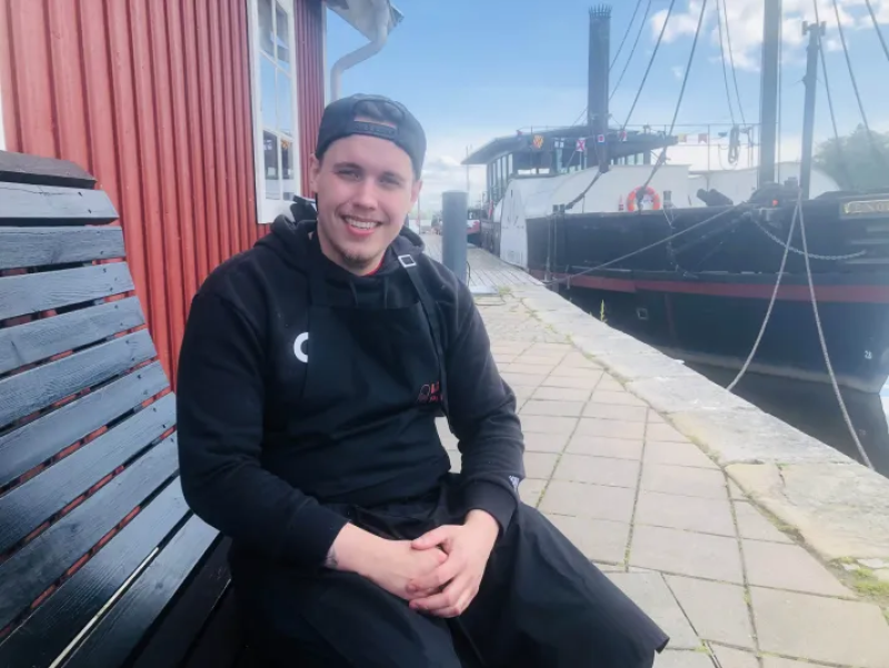 En man i svarta kläder och keps sitter på en bänk bredvid en röd byggnad nära en båt vid kaj en solig dag.