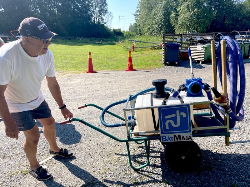 En man i vit skjorta och keps står bredvid en bärbar pump på hjul märkt 