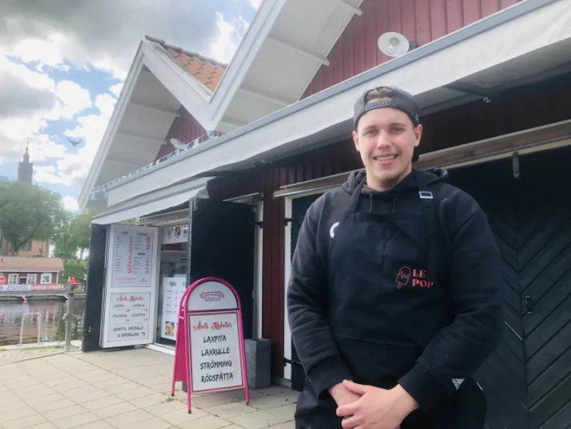 En man i ett svart förkläde står utanför en matkiosk med en menyskylt, bredvid en röd träbyggnad under en molnig himmel.