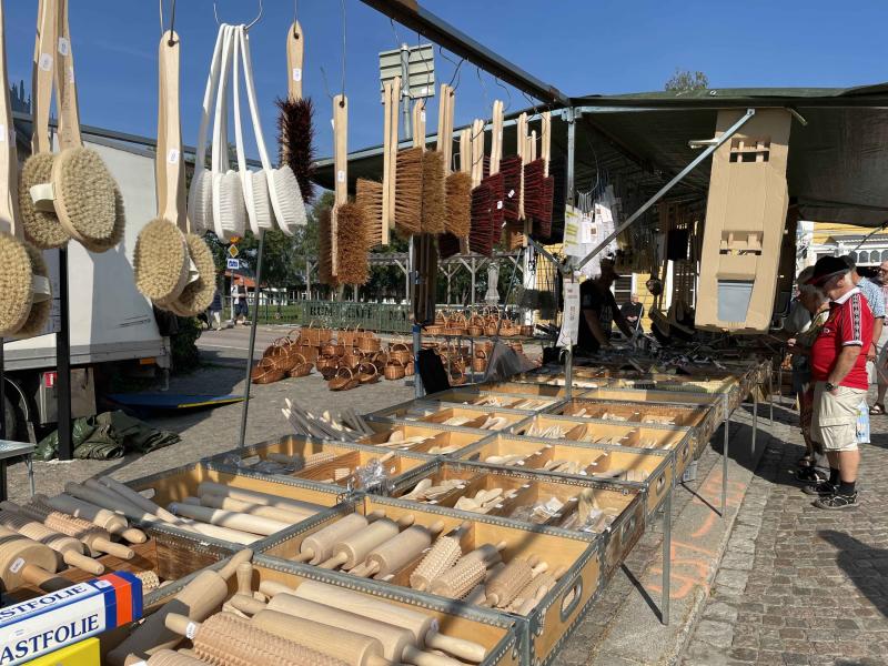 Ett marknadsstånd visar en mängd olika hängande penslar och kvastar, tillsammans med rader av kavel, köksredskap i trä och skärbrädor under ett tak en solig dag.