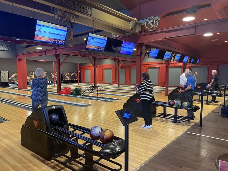 Människor som bowlar på en inomhusgränd med flera banor och överliggande skärmar. Bowlingklot och ställ är synliga.