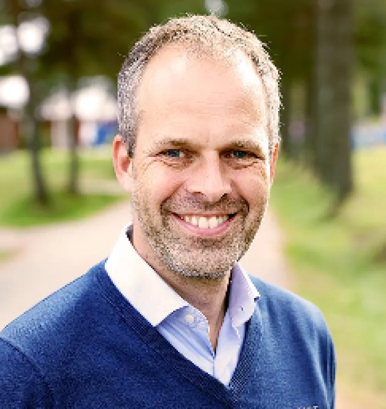 En man med kort hår och skägg ler medan han står utomhus på en stig med grönska i bakgrunden. Han är klädd i en blå tröja över en skjorta med vit krage.