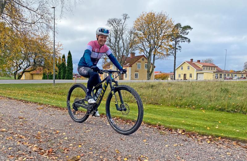 En cyklist i rosa och blå tröja cyklar på en mountainbike på en grusväg med hus, träd och en molnig himmel i bakgrunden.