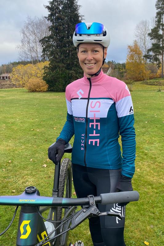 En cyklist med hjälm, solglasögon och en rosa och blå cykeltröja står utomhus bredvid en mountainbike på en gräsmatta med träd i bakgrunden.