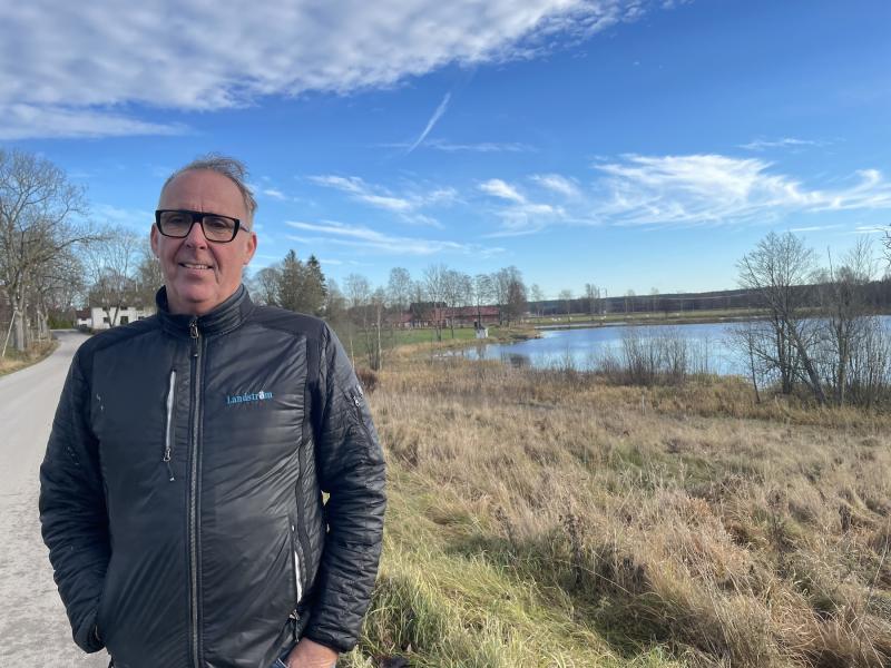 En man i svart jacka står på en landsväg bredvid ett gräsbevuxet landskap med en damm under en blå himmel.