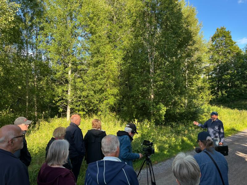 En grupp människor lyssnar på en person som talar nära ett skogsområde en solig dag, medan en person filmar scenen med en kamera på ett stativ.