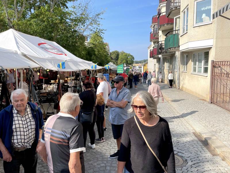 Människor går längs en marknadsgata kantad av försäljarstånd och hyreshus en solig dag.