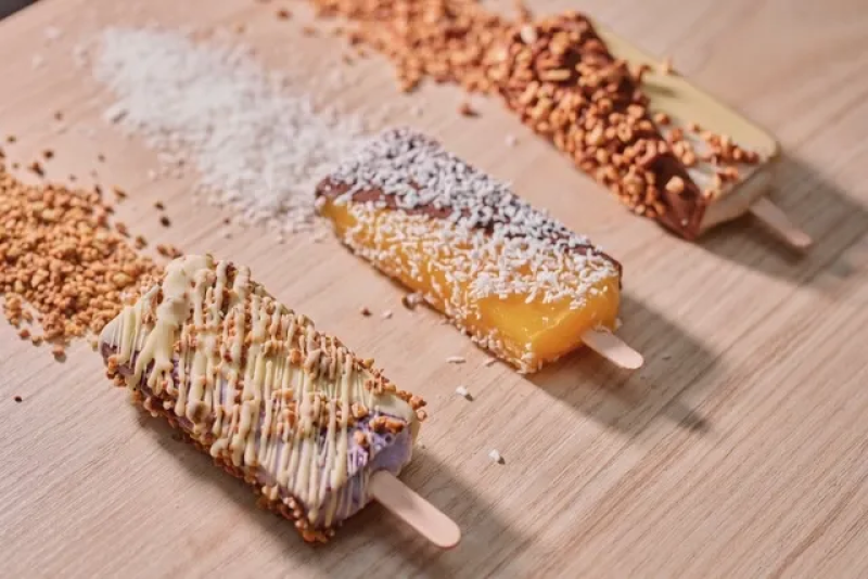 Tre isglassar på en träyta, var och en toppad med kokosflingor eller krossade nötter, med matchande toppings utspridda runtom.