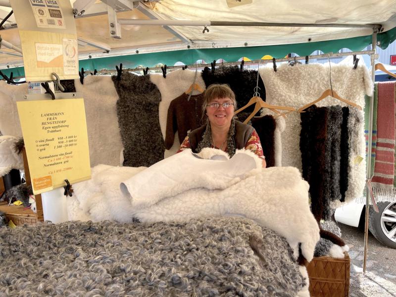 En person står bakom ett stånd och visar olika ylle- och pälsprodukter på en marknad. En gul skylt med produktdetaljer syns till vänster.
