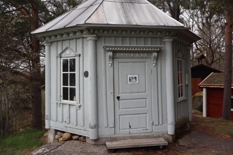 Liten, åttkantig, grå träbyggnad med plåttak, vitkantade fönster och en ytterdörr, omgiven av träd och andra träkonstruktioner.