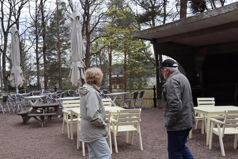 Två personer i jackor går förbi tomma utomhusbord och stolar på ett kafé eller en restaurang, med träd och stängda parasoller i bakgrunden.