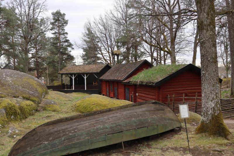 Flera traditionella träbyggnader med röda väggar och grästak ligger bland träd och mossiga stenar, med en gammal träbåt i förgrunden.