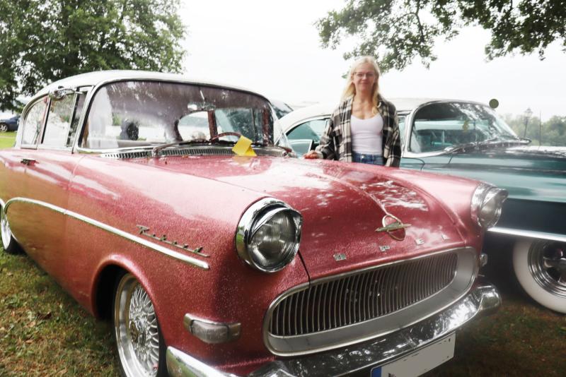 En person står bredvid en röd Opel Rekord från förr som står parkerad på gräset, och i bakgrunden syns en annan klassisk bil.