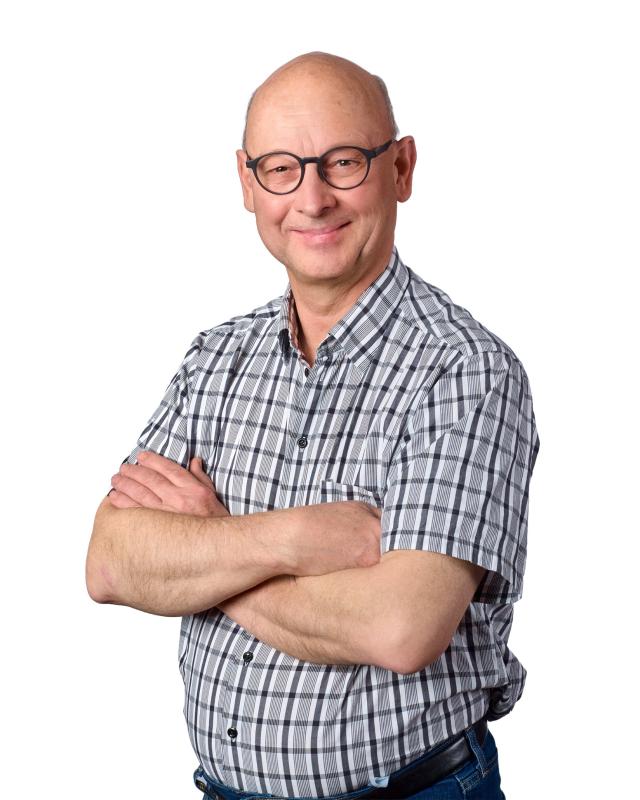 En skallig man med glasögon, klädd i en rutig kortärmad skjorta, står med armarna i kors och ler framför en vit bakgrund.
