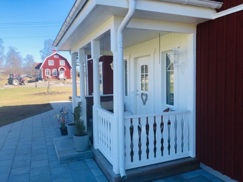 En liten vit veranda med krukväxter och en hjärtdekoration på dörren, kopplad till ett rött hus; en röd lada och en traktor syns i bakgrunden.