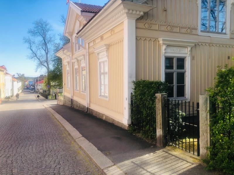 En lugn, solbelyst gata med ett gult trähus med vita lister och utsmyckade detaljer, intill en liten gated entré och kullerstensbelagd trottoar.