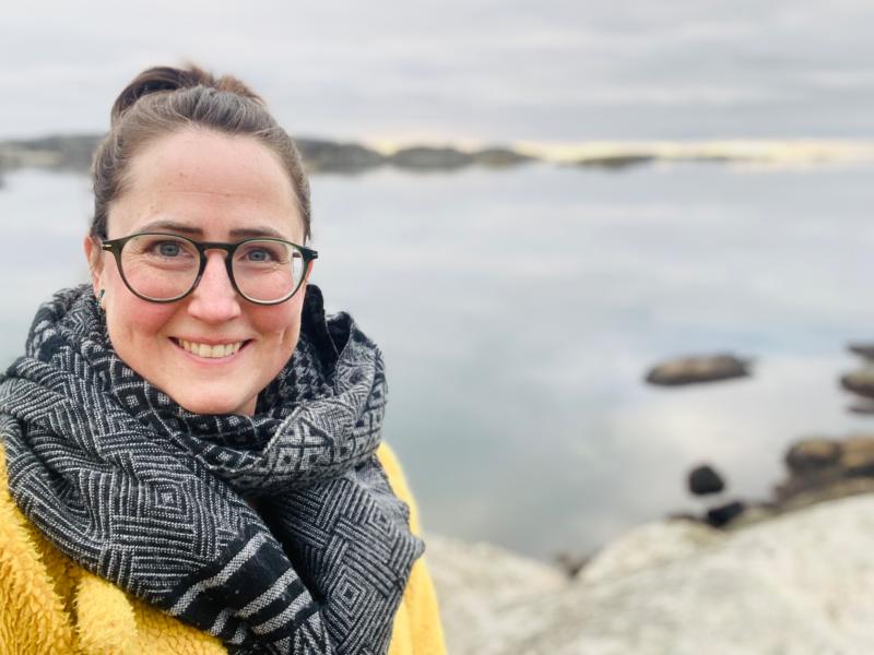 Kvinna med glasögon, mönstrad halsduk och gul kappa ler mot kameran med en klippig strand och lugnt vatten i bakgrunden under en mulen himmel.