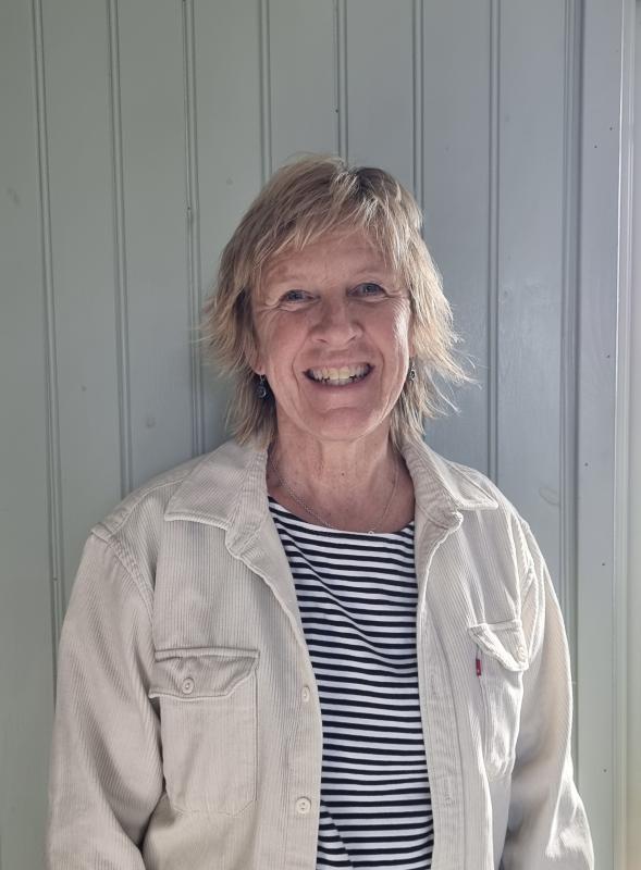 En kvinna med kort blont hår, klädd i en ljus jacka över en svartvitrandig skjorta, står leende framför en ljus vertikal panelvägg.