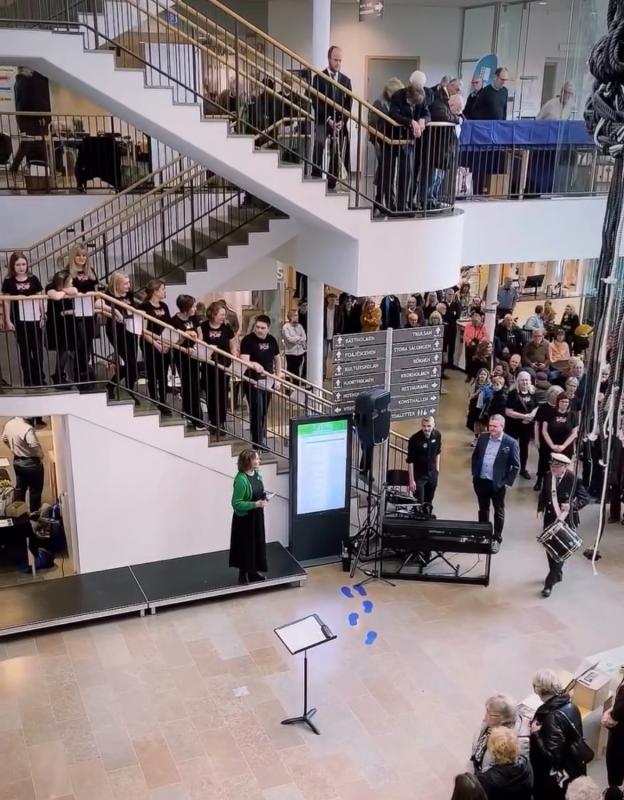 En kör står i en trappa och på en trappavsats och uppträder för en publik som samlats i en byggnad med flera våningar, glasräcken och informationsskyltar.