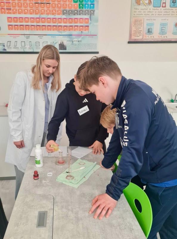 Tre studenter utför ett vetenskapligt experiment vid ett klassrumsbord medan en lärare i labbrock observerar. En affisch med periodiska systemet hänger på väggen bakom dem.