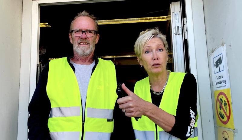 Två personer som bär gula säkerhetsvästar står i en dörröppning. Mannen till vänster har glasögon medan kvinnan till höger pekar fram och ser förvånad ut.