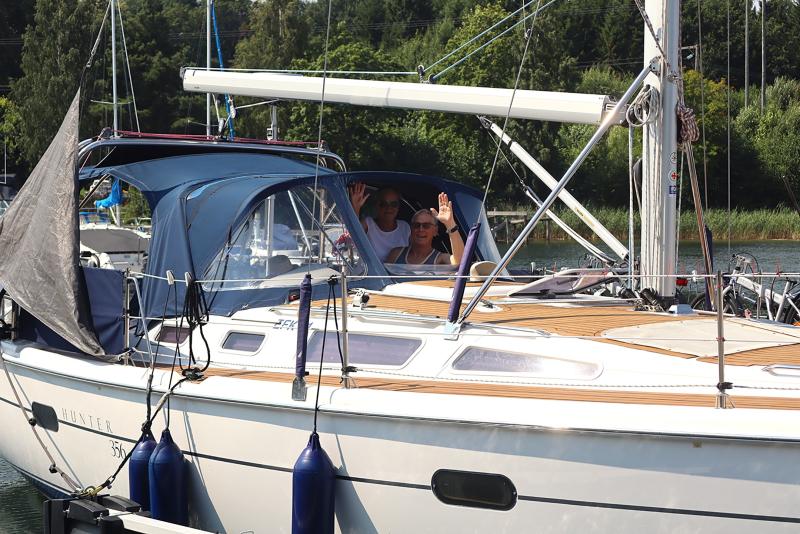 Två personer sitter på en förtöjd segelbåt och vinkar mot kameran. Segelbåten har ett blått tak och omges av vatten och grönska i bakgrunden.