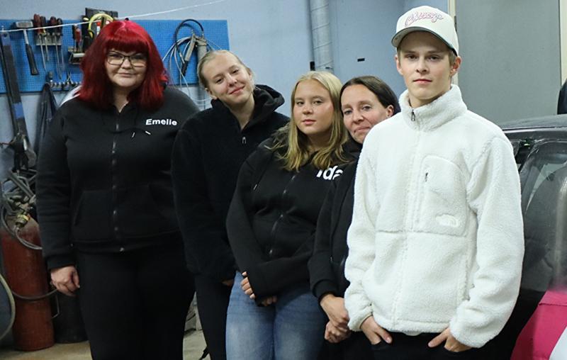 Fem personer står inomhus i en verkstad, klädda i vardags- eller arbetskläder, med verktyg och utrustning synliga i bakgrunden.
