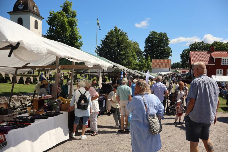 Folk tittar på varor vid marknadsstånden utomhus en solig dag, med en kyrka och träd i bakgrunden.